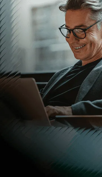 A man with glasses and a blazer smiles while he reads an article on a laptop