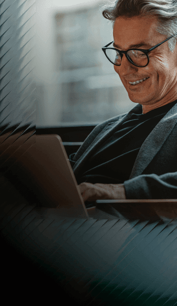 A man with glasses and a blazer smiles while he reads an article on a laptop