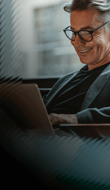 A man with glasses and a blazer smiles while he reads an article on a laptop