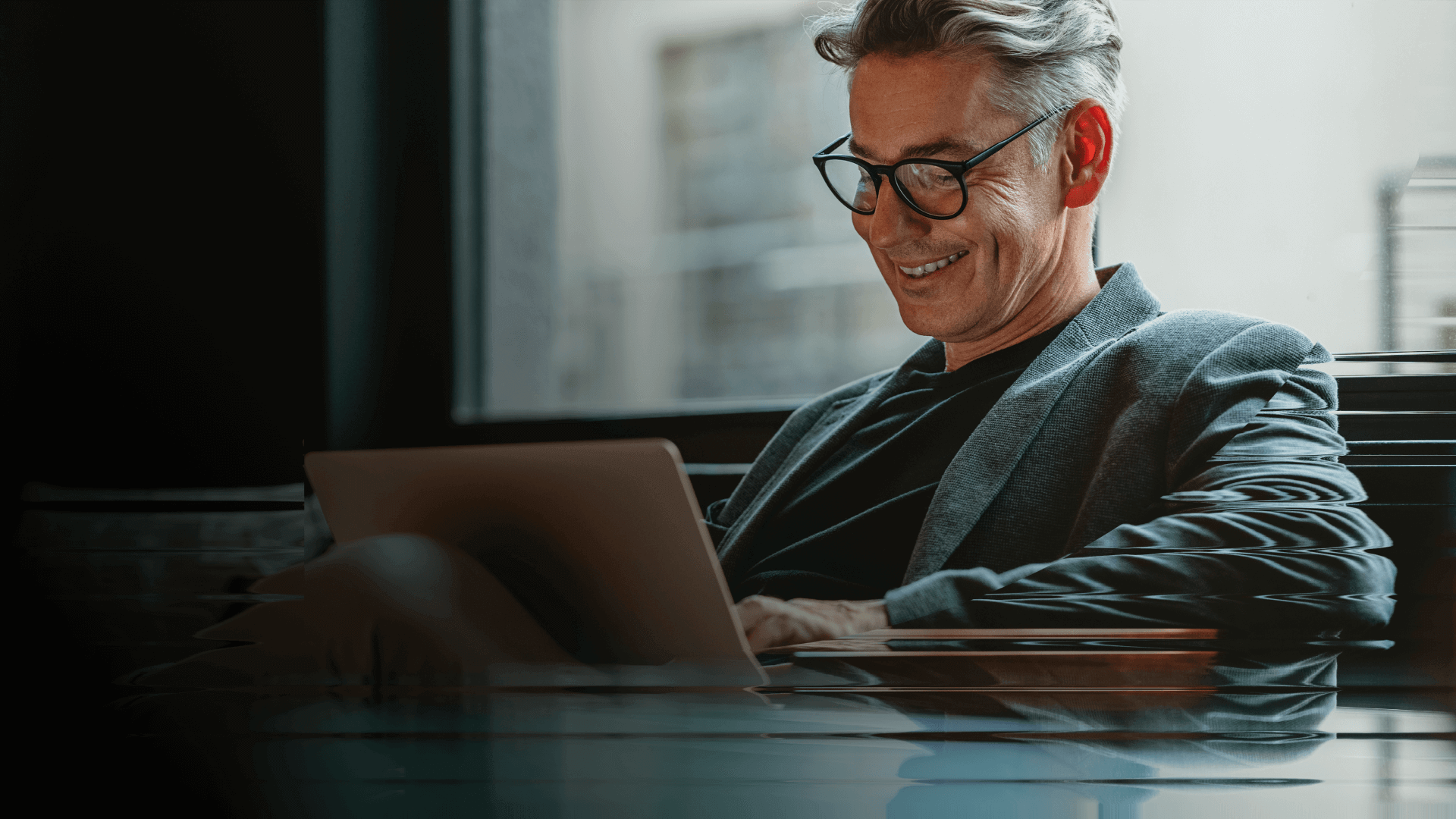 A man with glasses and a blazer smiles while he reads an article on a laptop