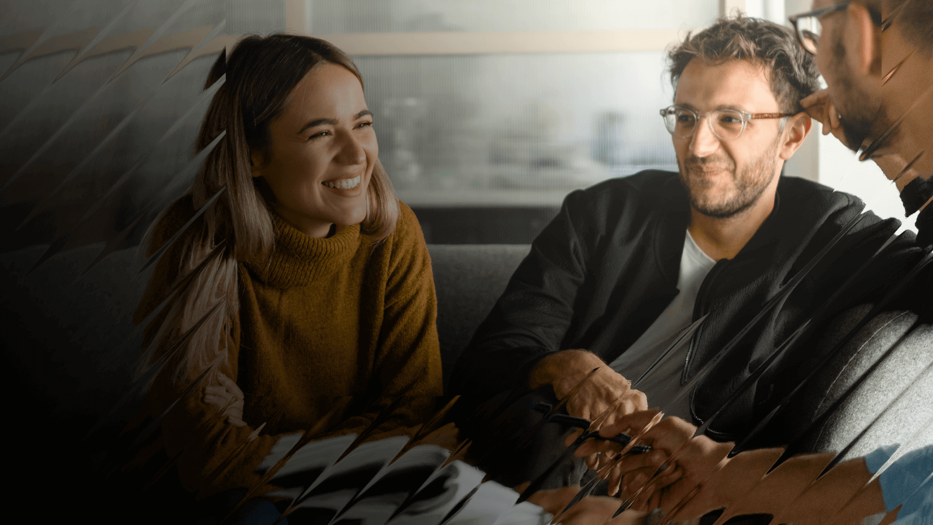 Three people sit in a casual meeting space, smiling while discussing something