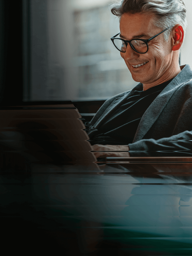 A man with glasses and a blazer smiles while he reads an article on a laptop